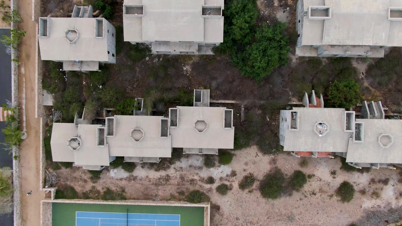 Overhead San Jose del Cabo view with pale flat roofs across the upper and middle frame, darker green trees to the right, scrubby sand below, and a tennis court corner visible at the bottom, no visible people.