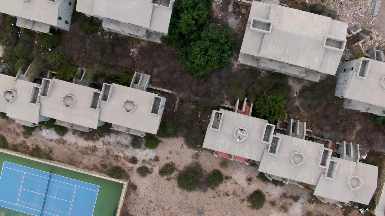 Overhead drone frame in San Jose del Cabo showing pale concrete rooftops, dry sandy ground, shrubs, trees, and the left edge of a blue-green tennis court, with no visible people.