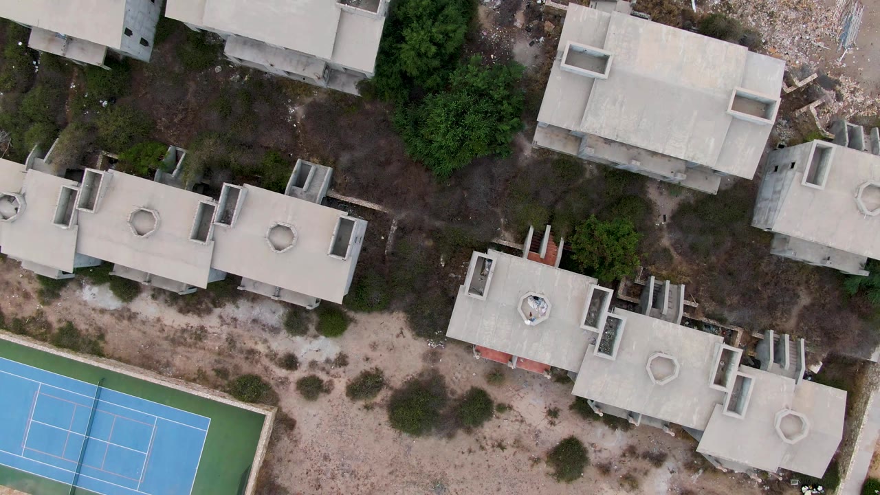 Concrete rooftops, trees, dirt paths, and a blue tennis court from above