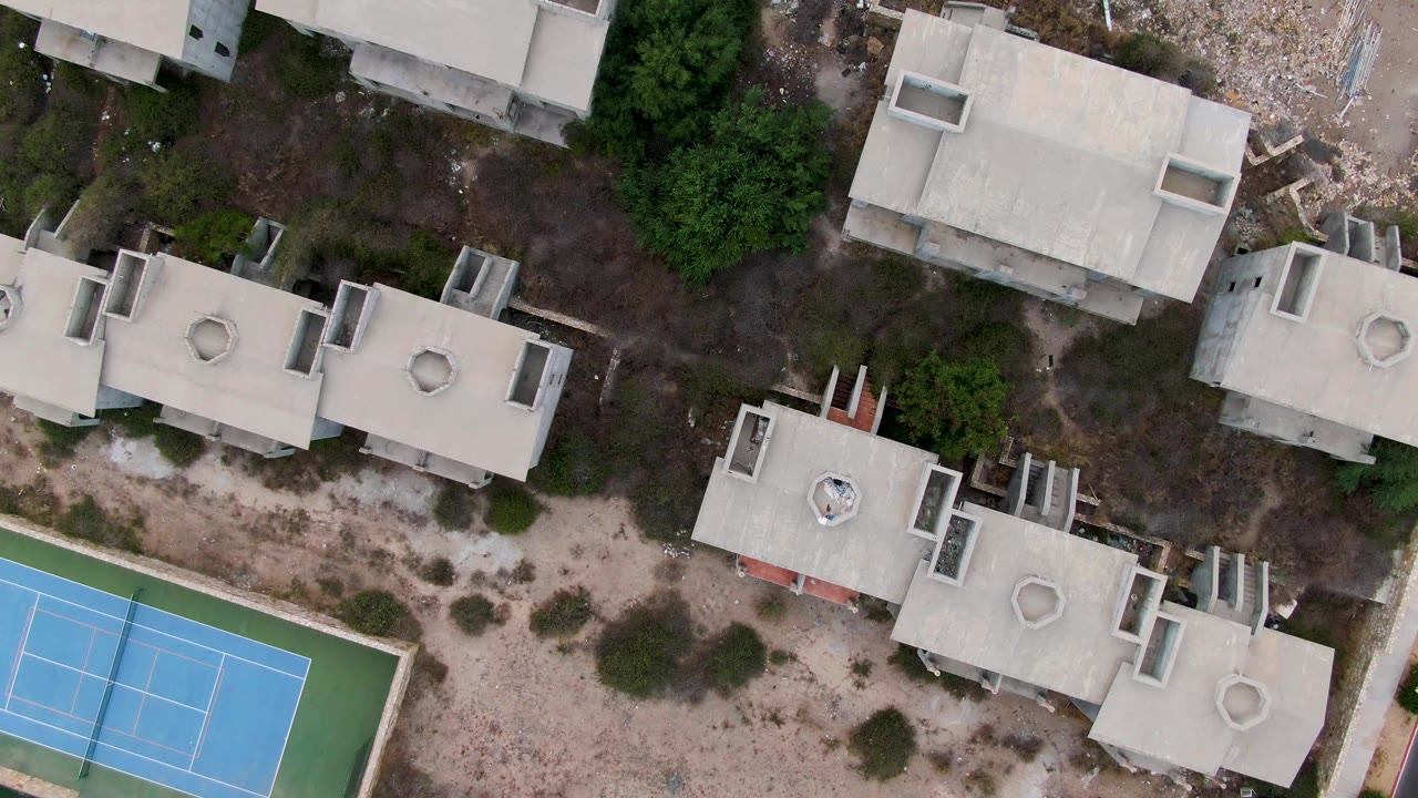 Straight-down aerial frame in San Jose del Cabo showing concrete residential roofs, a blue court with green perimeter, sandy ground, and dense trees along the upper area, with no visible people.