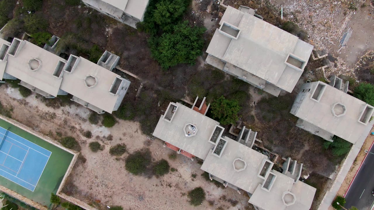 Overhead frame in San Jose del Cabo, Mexico showing flat-roof residential buildings, scrubby dirt lots, trees, and part of a blue tennis court with no visible people.