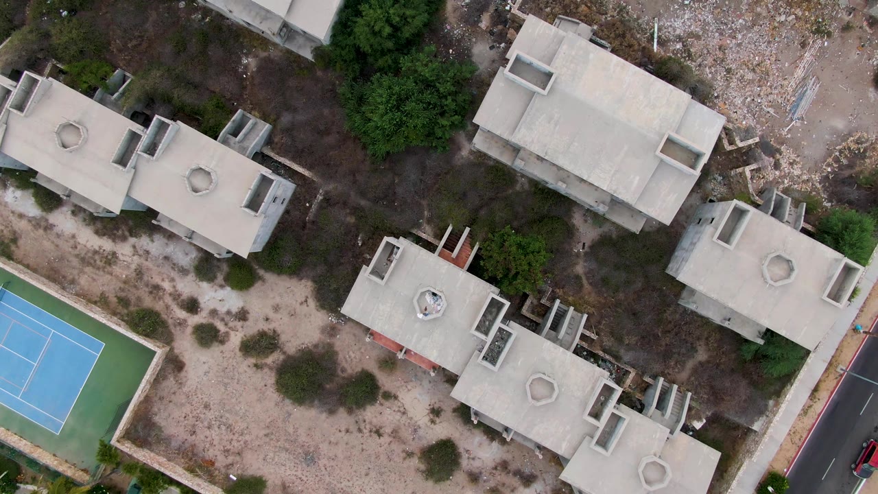 Aerial view of residential rooftops, scrub land, tennis court, and road edge.
