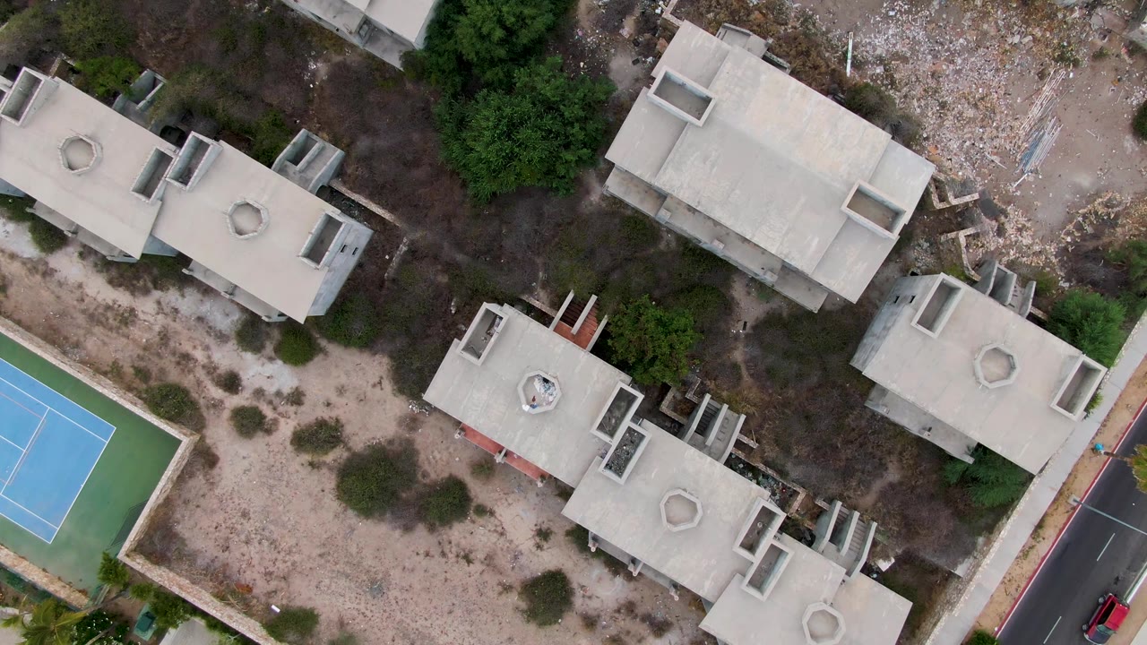 Aerial frame over San Jose del Cabo, Mexico showing clustered rooftops, dry lots, the tennis court, and a red car visible on the road at the right edge, with no visible people.