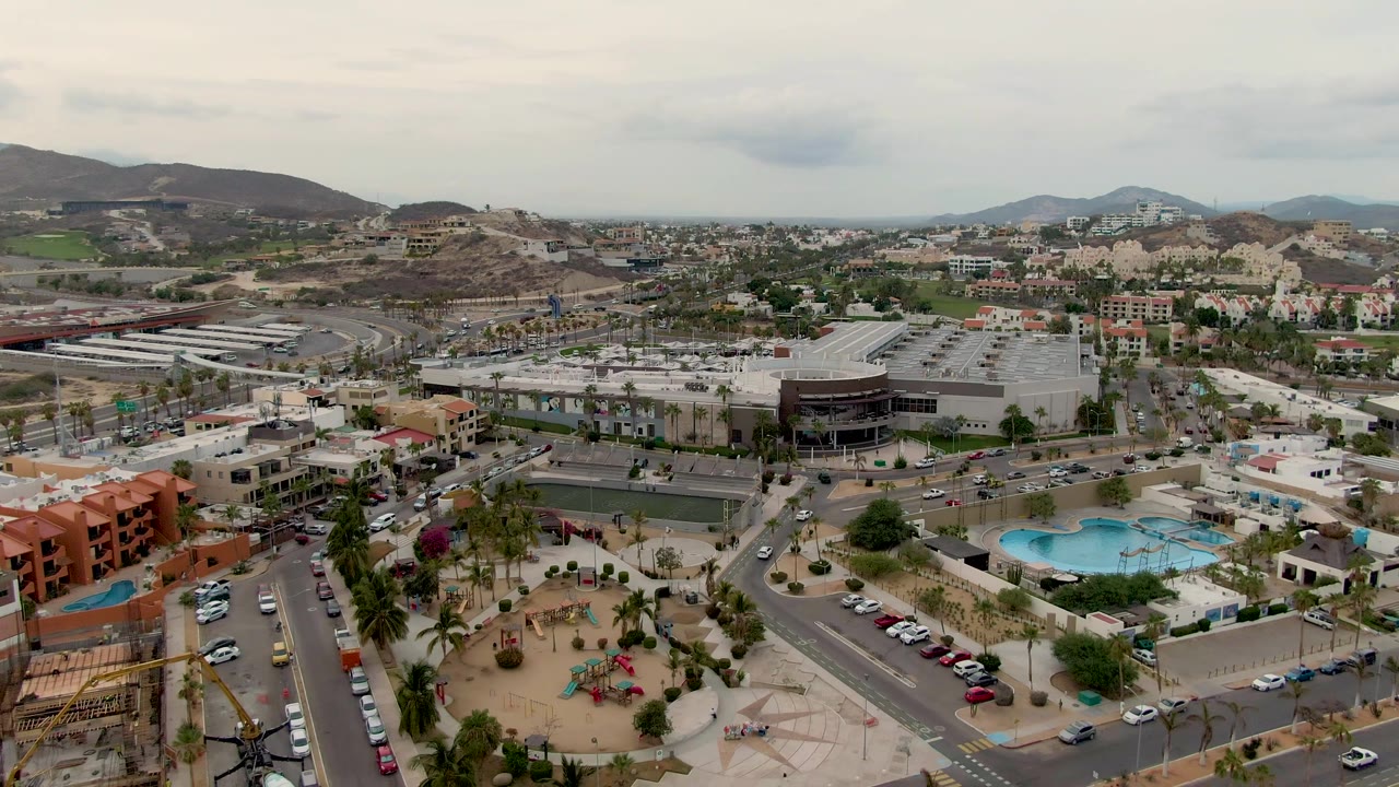 Aerial view across San Jose Del Cabo with orange residential buildings in the foreground, a pool complex, roads with cars, large parking lots, distant mountains, and no visible people.