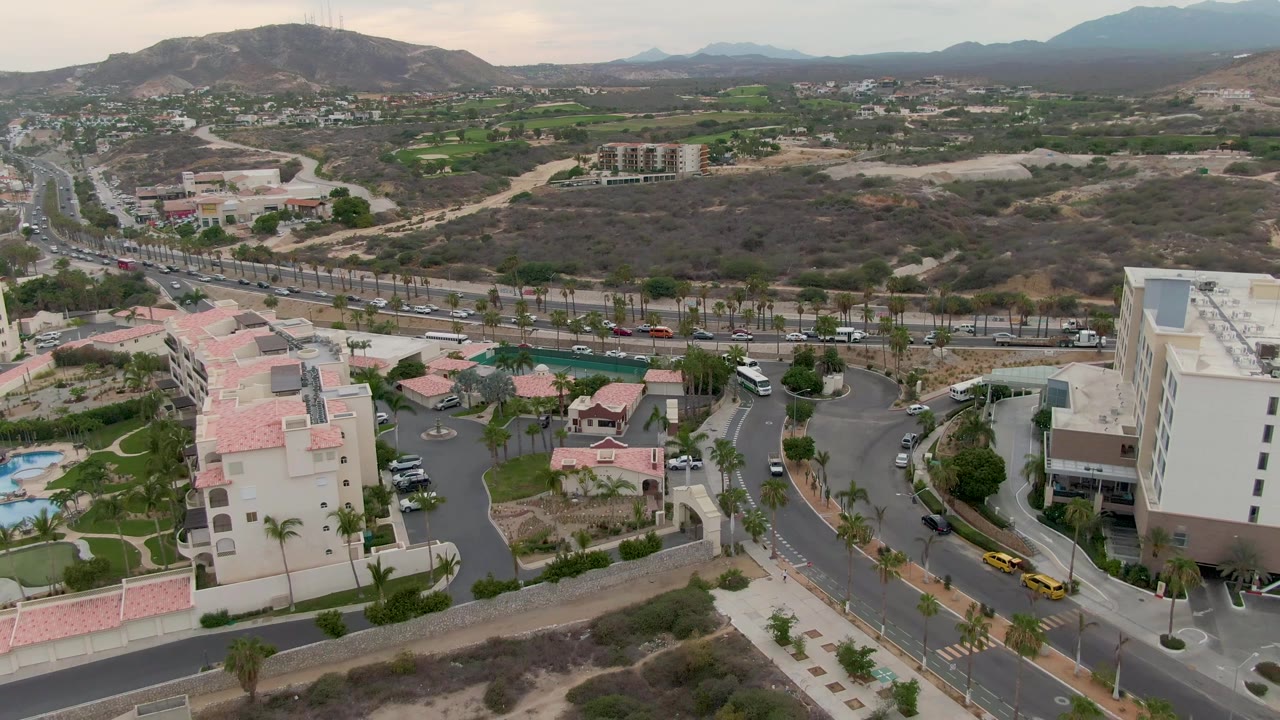 Opening aerial frame over San Jose del Cabo shows curving roads, pale buildings with red tile roofs, blue pools, dry lots, green patches, distant hills, and no visible people.