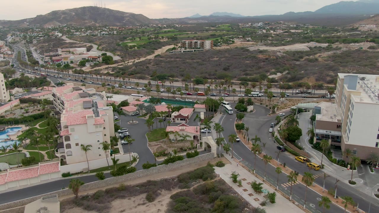 Aerial frame over San Jose del Cabo shifts across apartment and commercial buildings, palm-lined streets, moving traffic, dry scrubland, distant mountains, and no visible people.