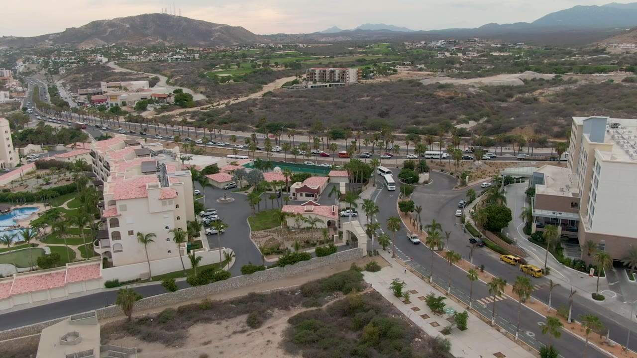 Mid-pan aerial frame in San Jose del Cabo shows a tall white building at right, red-roofed structures below, active roads, desert lots, green areas, hazy hills, and no visible people.