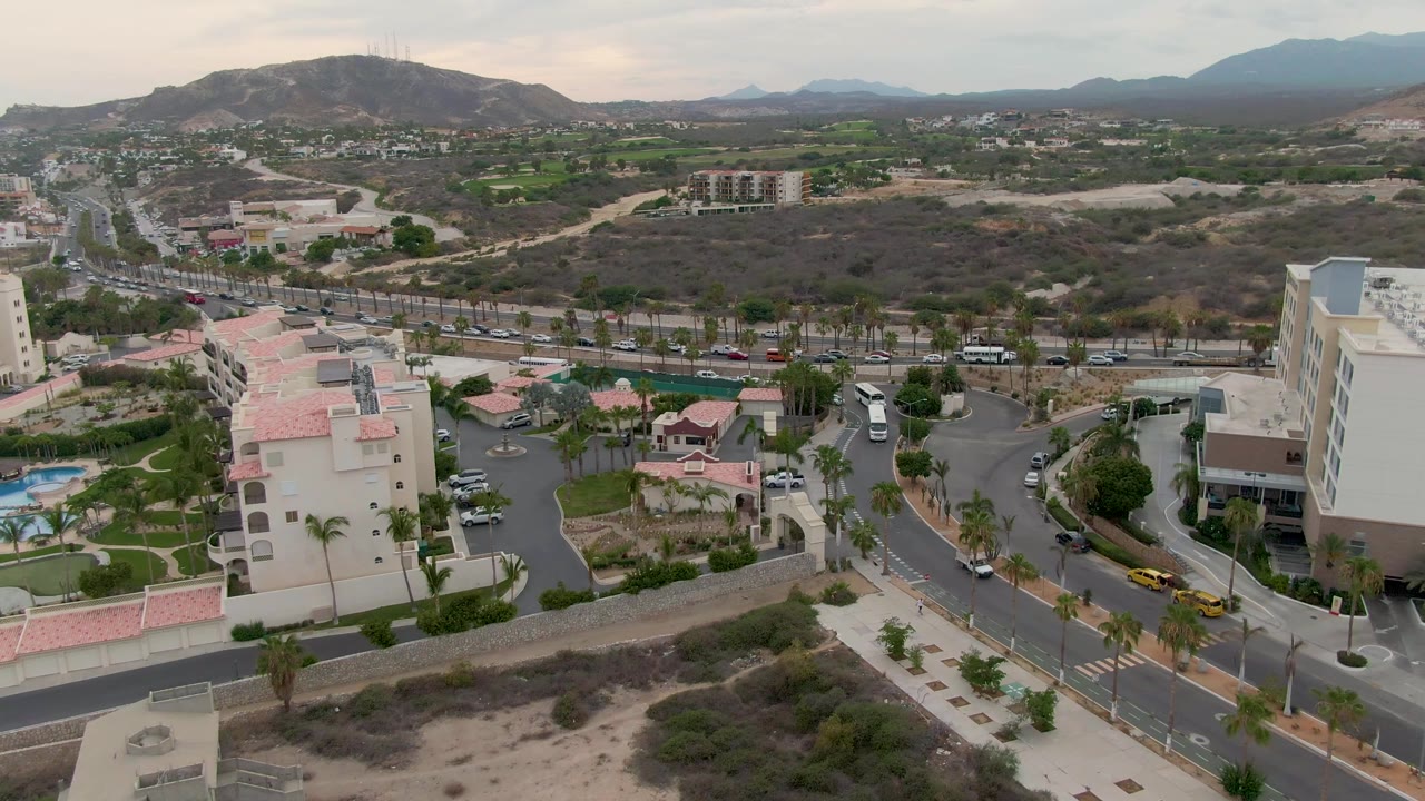Aerial neighborhood view with roads, vehicles, low buildings, pools, dry terrain, and mountains.