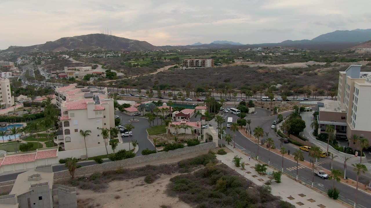 Opening frame of San Jose Del Cabo from above, with a curving road, pale buildings, palm trees, sandy scrub foreground, distant hills, cloudy sky, and no visible people.