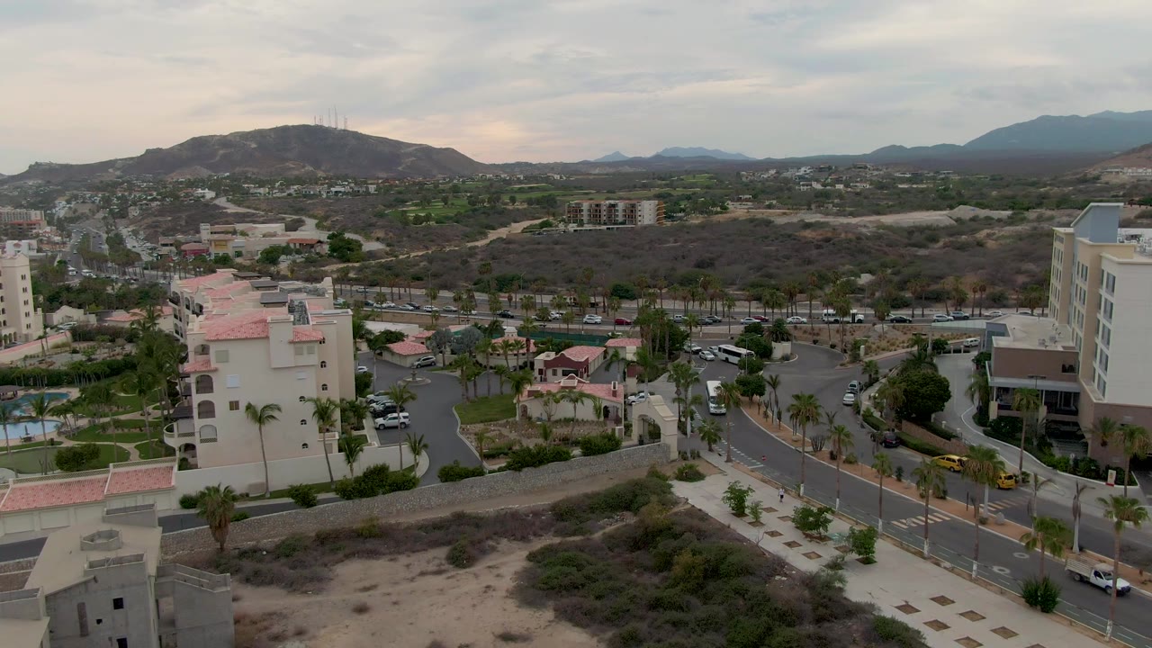 San Jose Del Cabo road scene one second in, showing vehicles along the street, hotel-like buildings, palms, dry open ground, layered hills, cloud cover, and no visible people.