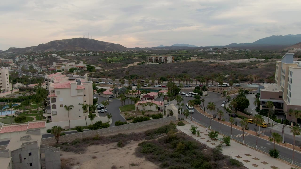 Mid-clip San Jose Del Cabo frame with a bus and cars on the road, cream-colored buildings on both sides, palms, sandy foreground, mountain silhouettes, cloudy sky, and no visible people.