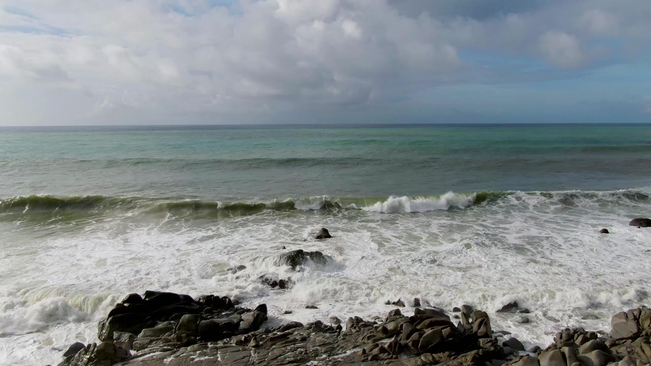 At Punta Gordo, Baja California Sur, white surf breaks across dark shoreline rocks with green-blue ocean and gray clouds beyond, no visible people.