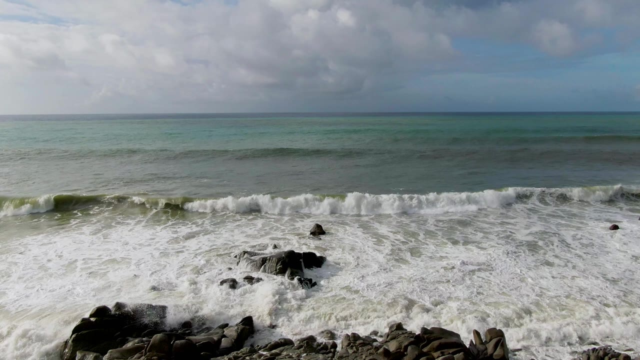At Punta Gordo, Baja California Sur, a thicker band of white foam pushes toward scattered dark rocks below a level ocean horizon, no visible people.