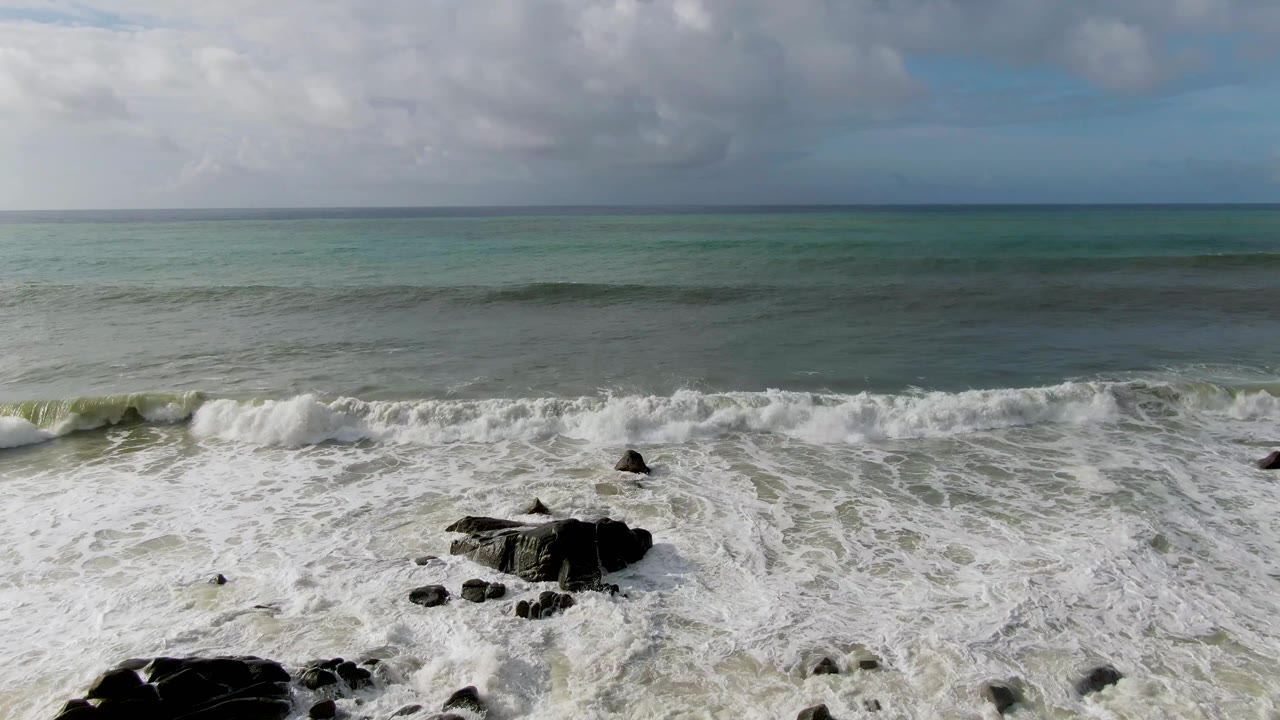 At Punta Gordo, Baja California Sur, whitewater expands across the lower frame around several exposed rock outcrops with darker water bands offshore, no visible people.