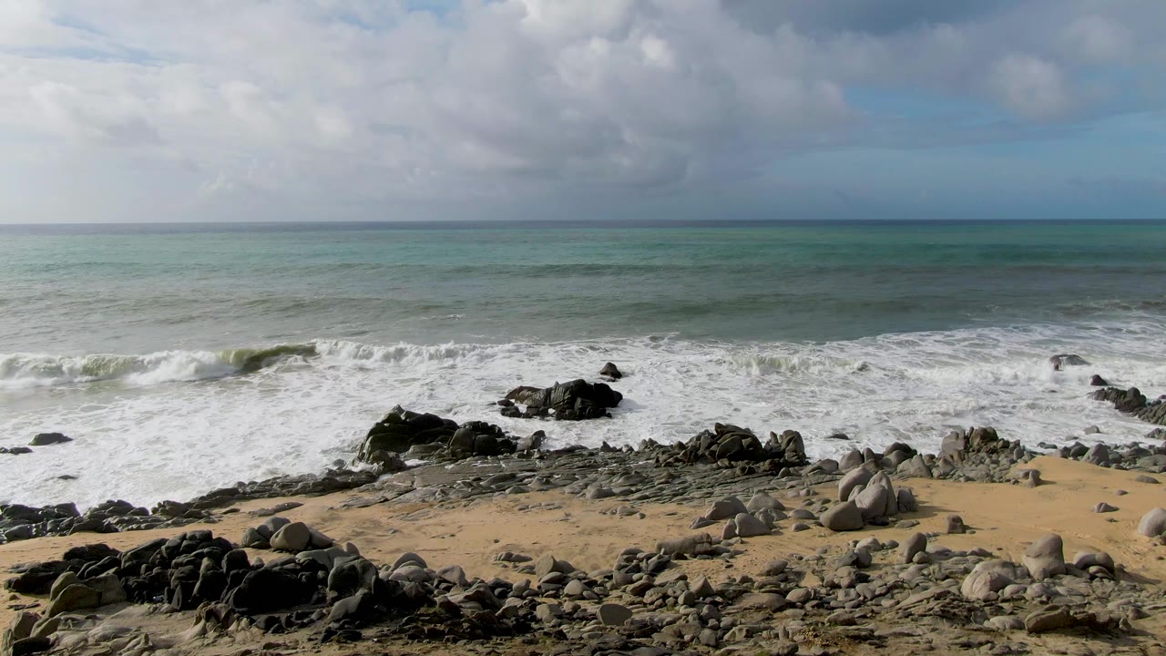 Wide view of Punta Gorda East Cape shoreline with tan sand, dark rocks, foamy surf, turquoise water, a cloudy sky, and no visible people.