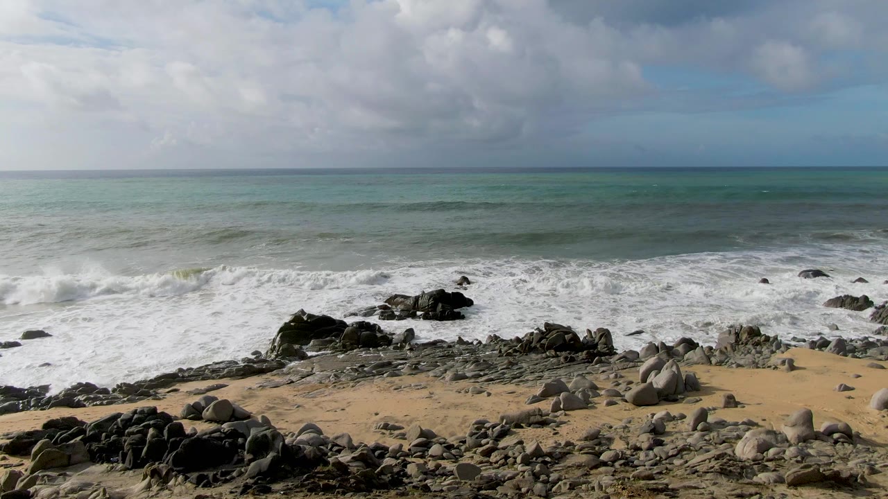 Punta Gorda East Cape surf pushes toward dark foreground rocks and pale sand, with white foam spread across the midground and no visible people.