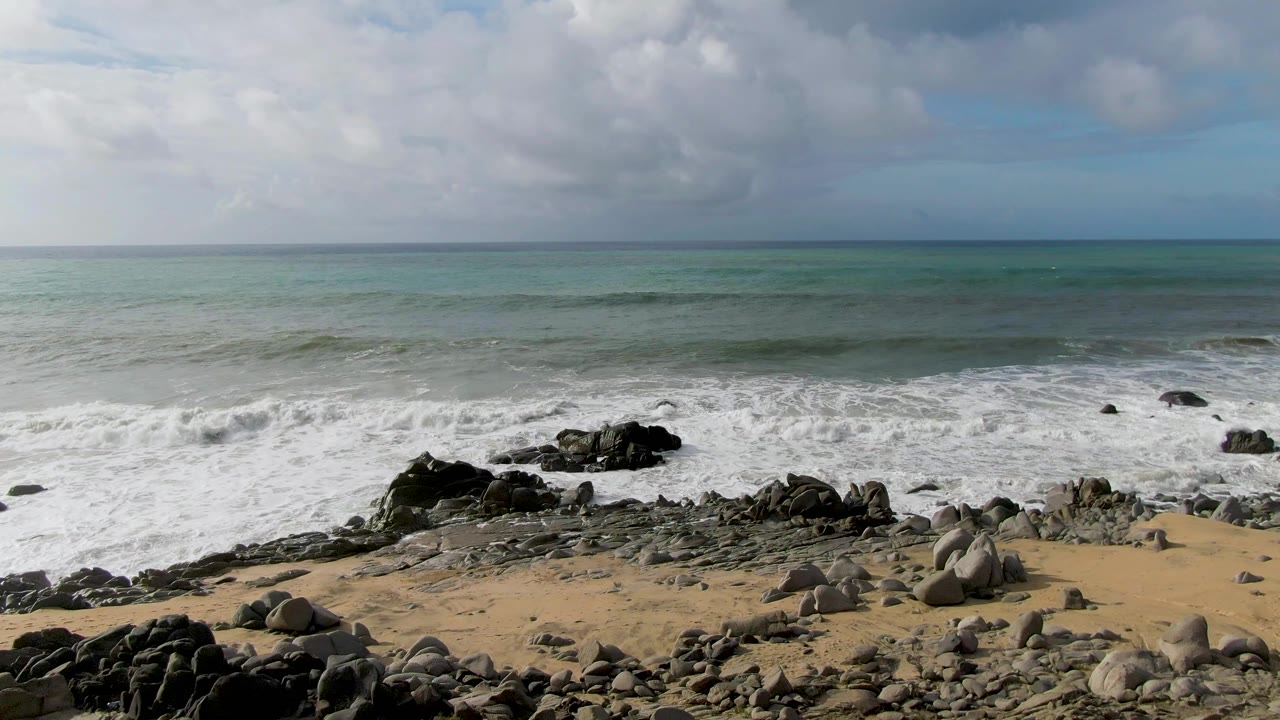A broad band of white surf covers the rocky shoreline at Punta Gorda East Cape, with scattered boulders on tan sand and no visible people.