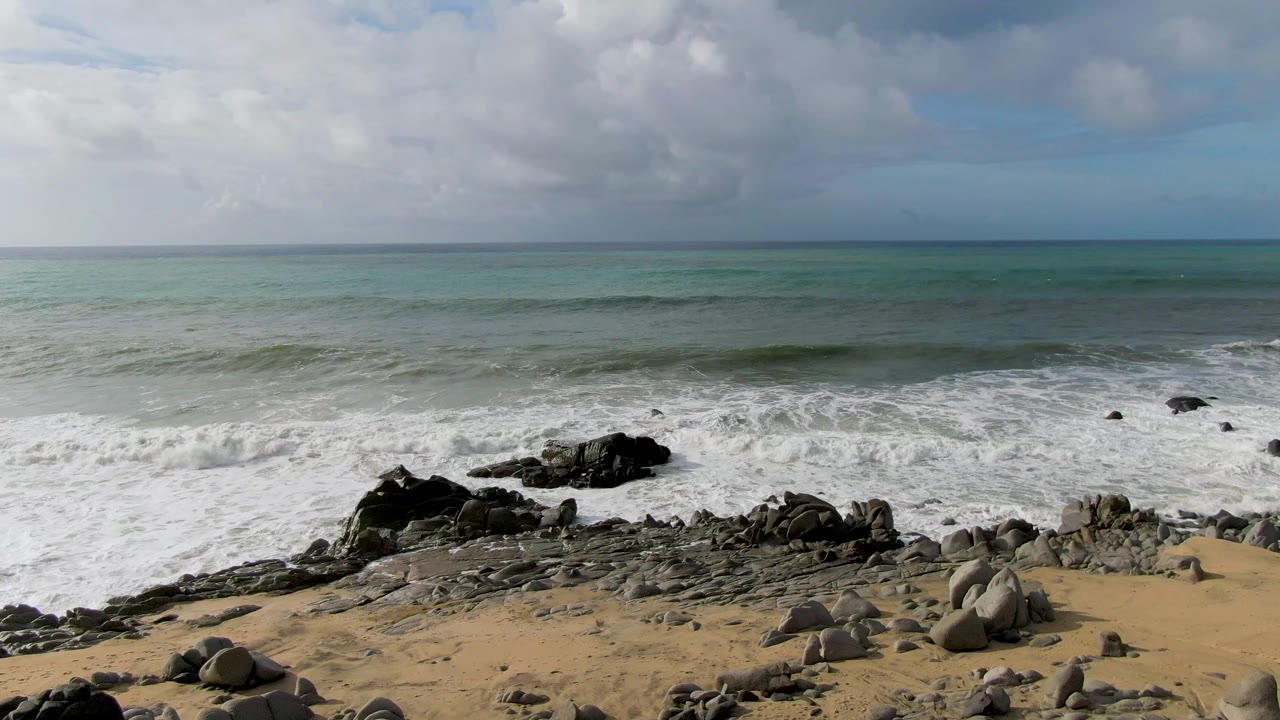 A larger wave line rolls in from the green-blue ocean toward black rocks and sandy beach at Punta Gorda East Cape, with no visible people.