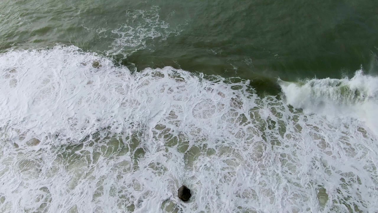Aerial frame from Punta Gorda, Baja California Sur, with a wide white foam band crossing green water and a small dark rock near the lower center, no visible people.