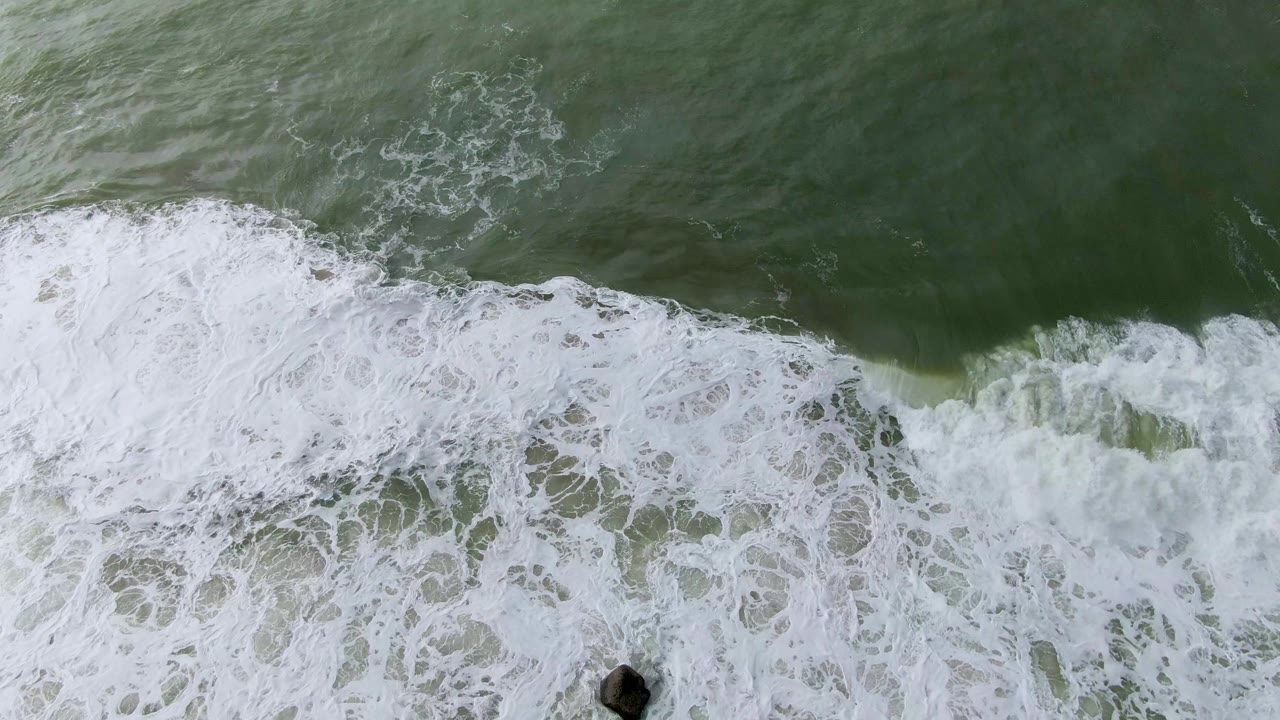 Top-down Punta Gorda surf frame with white foam curling across green water and a dark rock near the lower right edge, with no visible people.