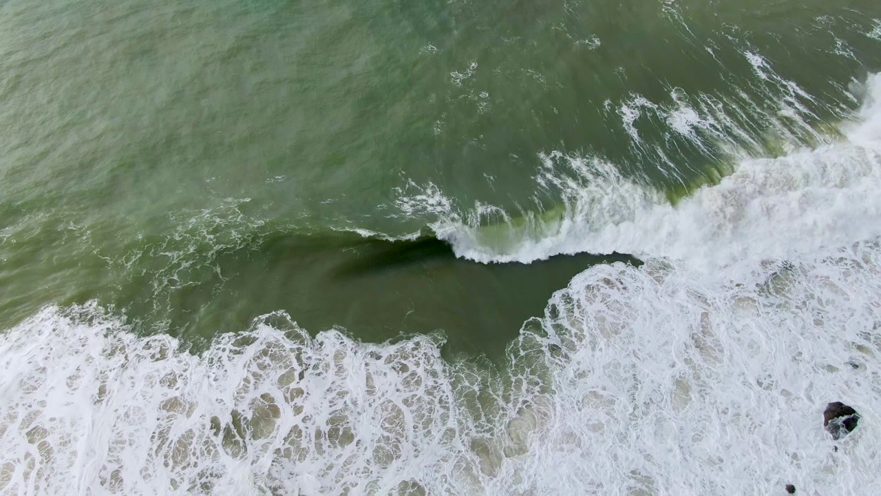 Straight-down view in Punta Gorda, Baja California Sur, showing white surf foam below a green breaking wave, with a small dark rock near the lower right and no visible people.