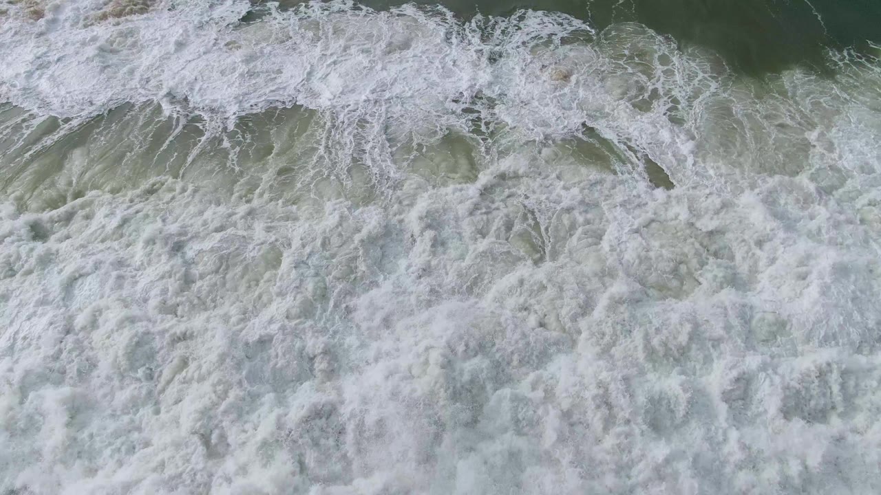 Top-down view of lace-like white foam covering most of the frame as a dark rock sits beneath the surf at Punta Gorda, Baja California Sur, with no visible people.