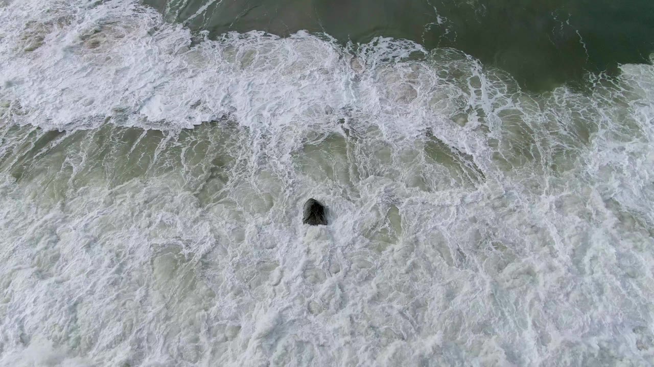 Aerial ocean frame with a broad white foamy wave crossing green water and one dark rock visible on the right side at Punta Gorda, Baja California Sur, with no visible people.