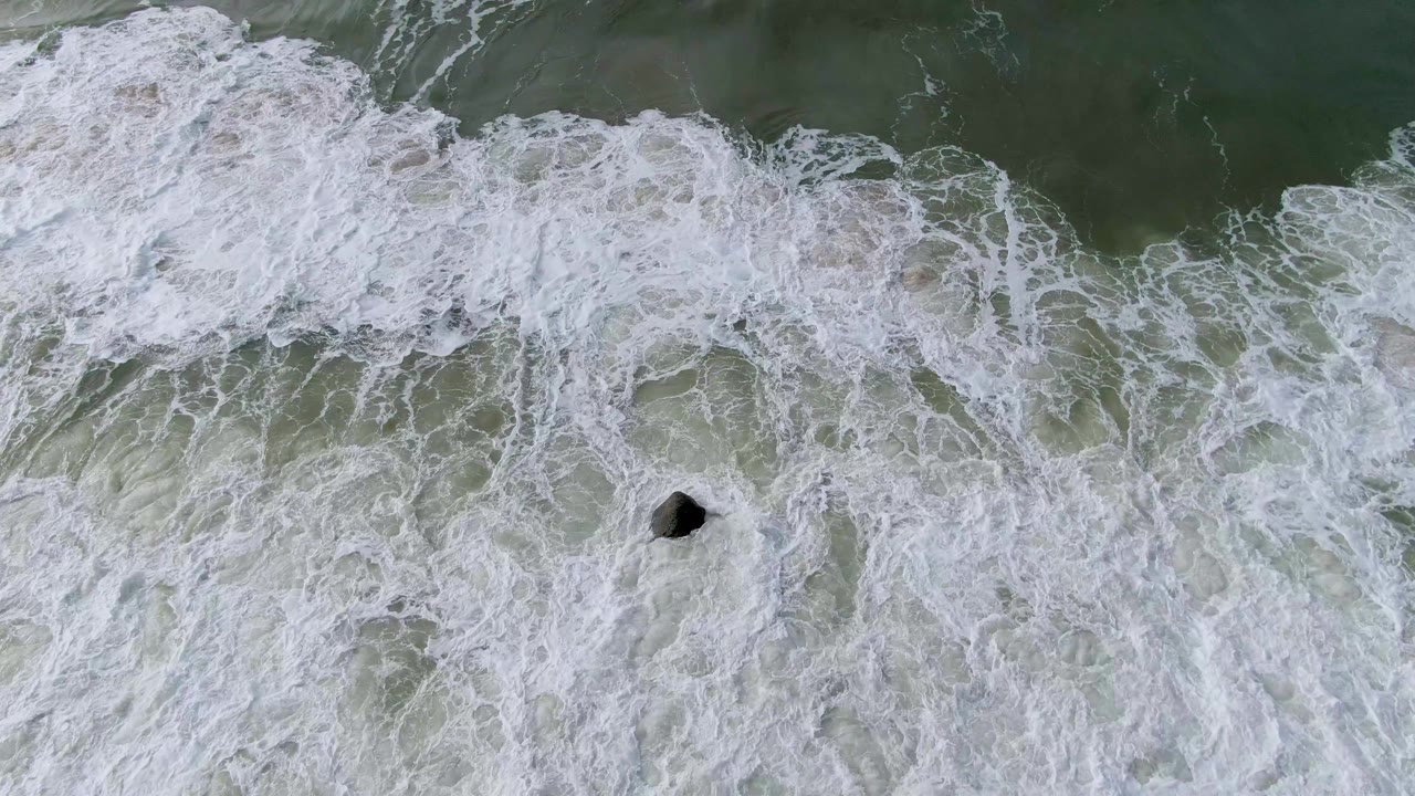 Overhead view of receding white foam around a dark submerged rock near the lower center of the ocean frame at Punta Gorda, Baja California Sur, with no visible people.