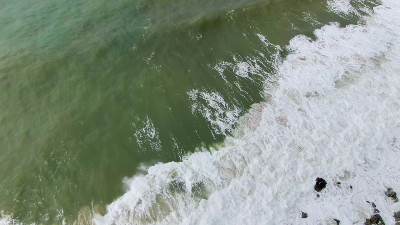 Overhead view in Punta Gorda, Baja California Sur, of green ocean water breaking into white foam across dark rocks near the lower right, with no visible people.