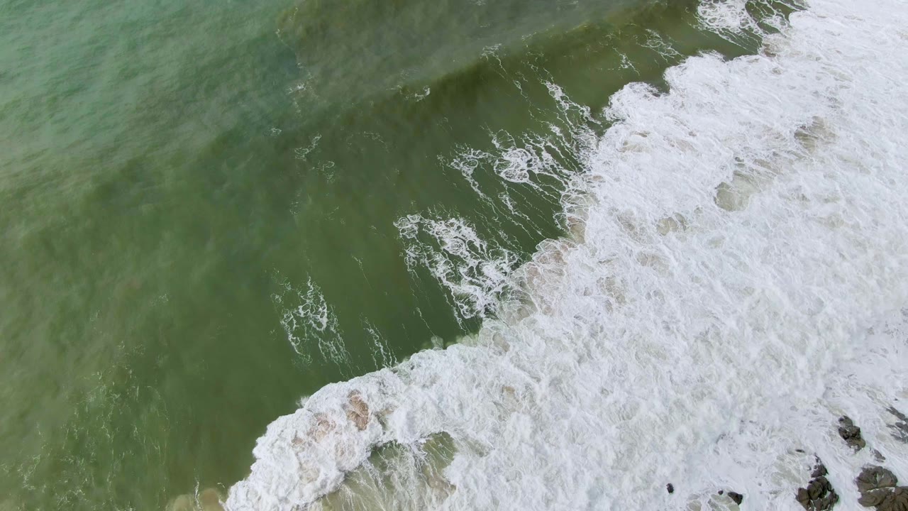 Overhead Punta Gorda coastal frame with a broad white foam band pushing diagonally through green water and small rocks visible near the right edge, with no visible people.