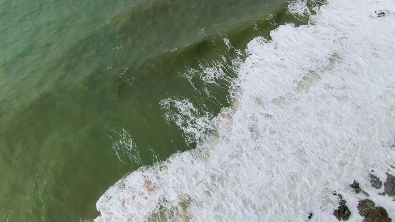 Overhead frame from Punta Gorda showing dense white surf foam sweeping across the lower and right side of green water, with dark rocks at the edge and no visible people.
