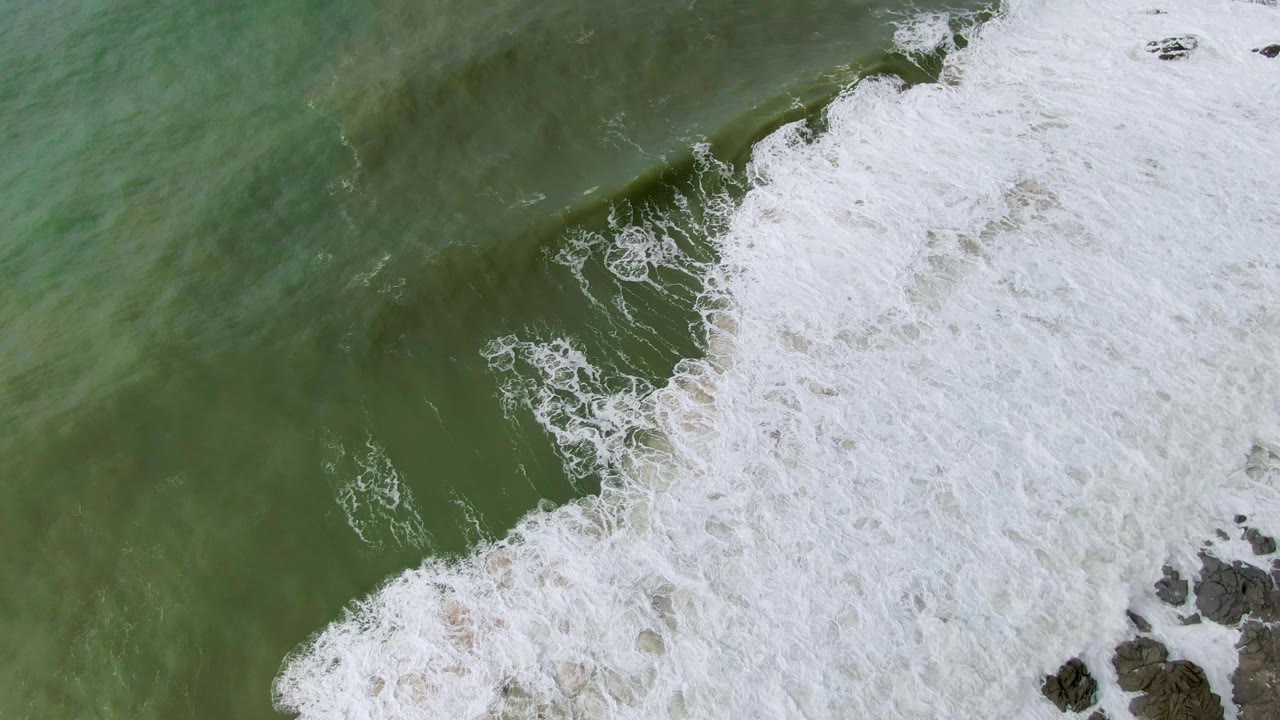 Large sheet of white foam spreads over green ocean and shoreline rocks.