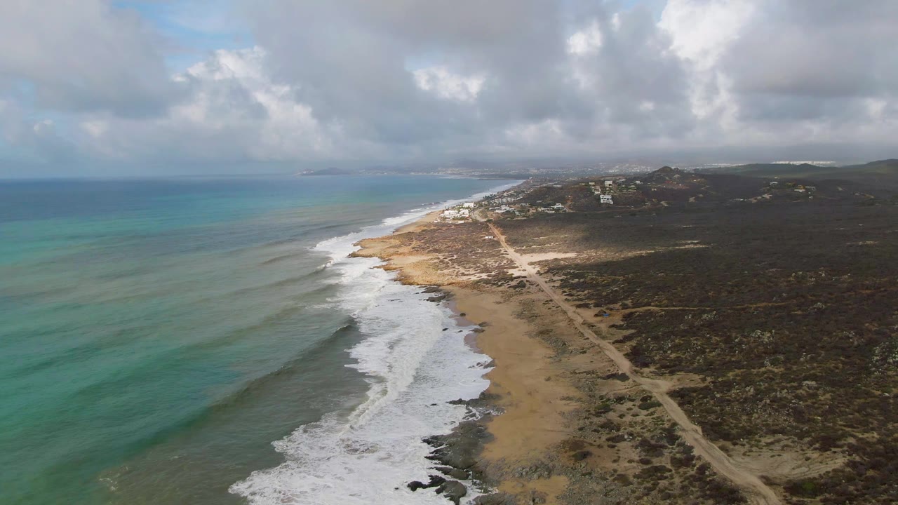 Aerial view over Punta Gorda, Baja California Sur, with turquoise ocean on the left, white surf along the sandy shore, a dirt coastal road through scrubland, and no visible people.