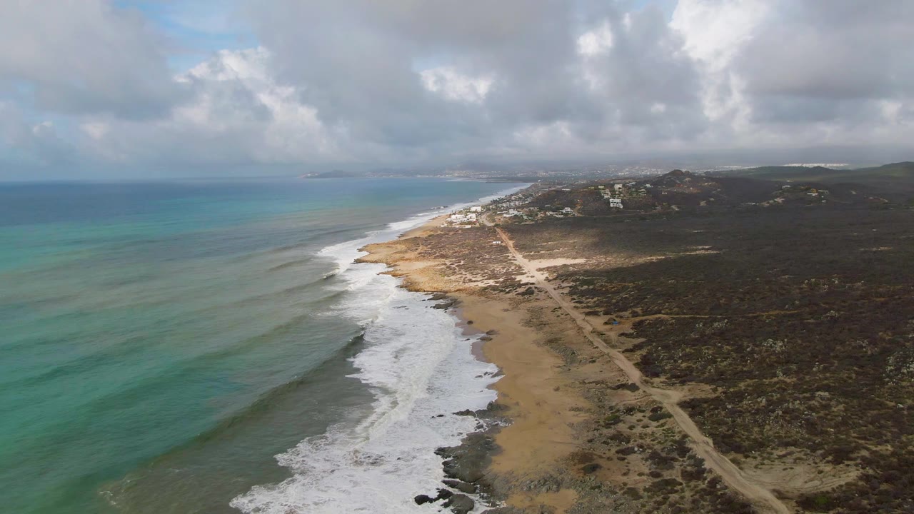 High aerial frame of Punta Gorda coastline showing a long band of white waves, tan beach, scrub-covered land, a pale road, distant buildings, and no visible people.
