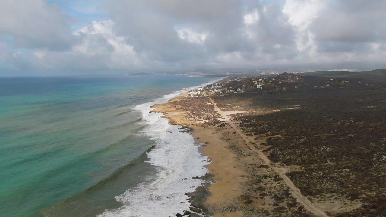 Drone view over Punta Gorda with green-blue water, foamy breakers, a narrow sandy shore, dark scrubland, small distant structures, cloud shadows, and no visible people.
