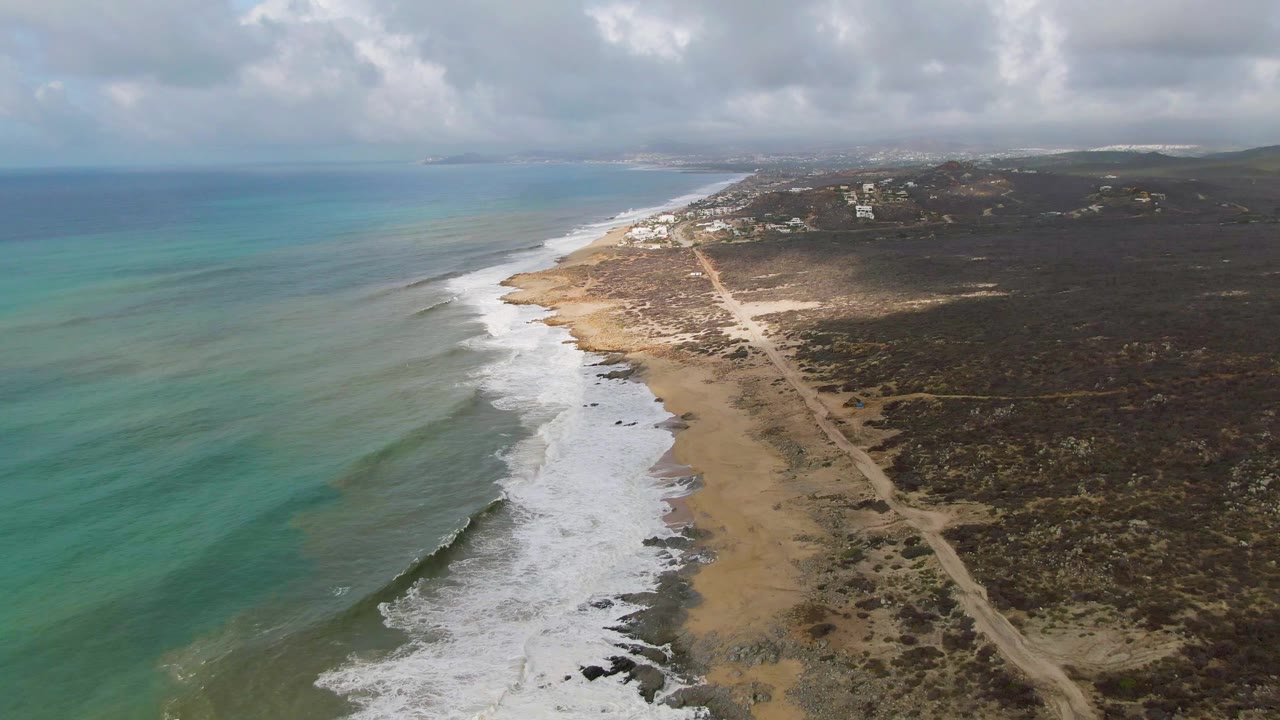High aerial view of Punta Gorda, Baja California Sur with turquoise water on the left, white surf on a sandy and rocky shoreline, a dirt road through scrub vegetation, and no visible people.