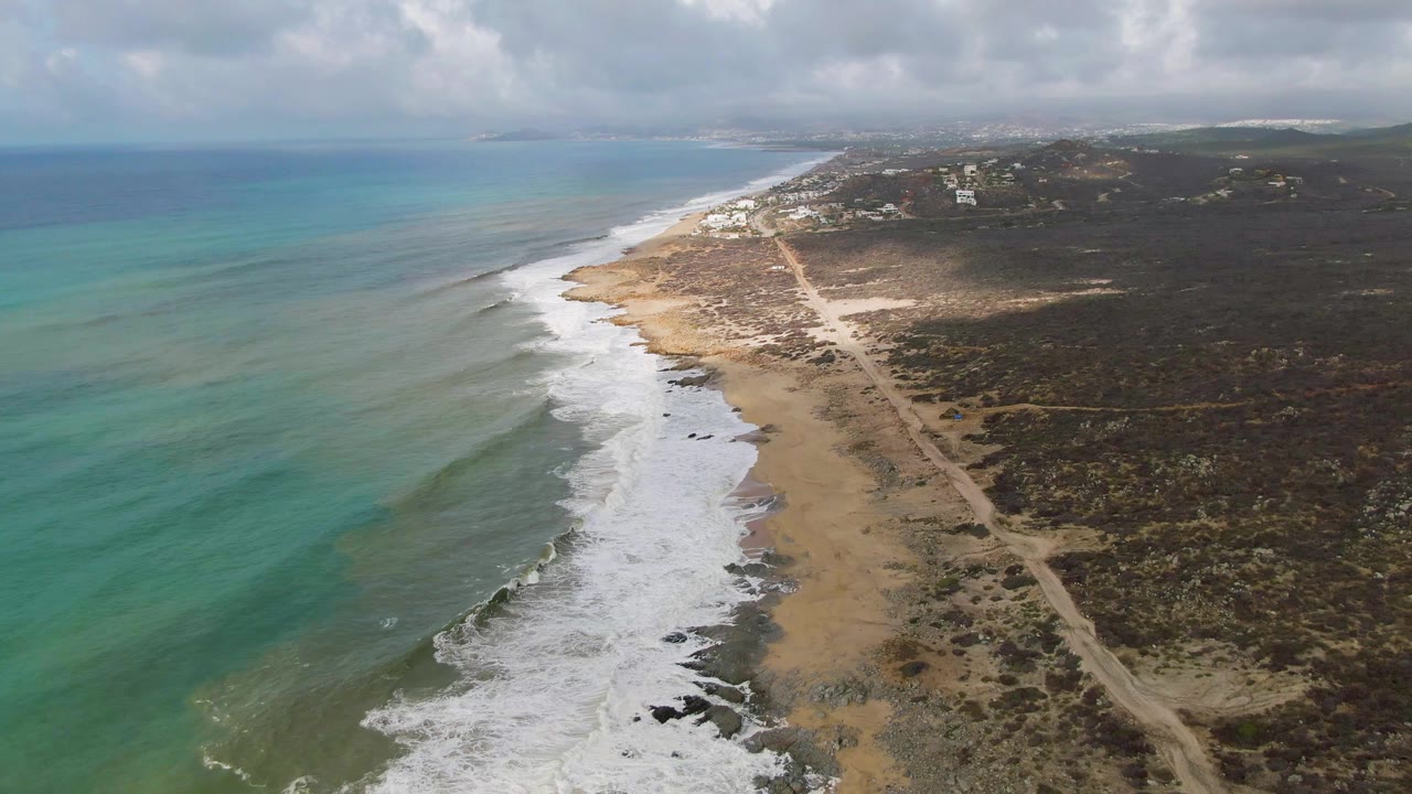 Aerial frame over Punta Gorda showing blue-green water with darker bands, a broad strip of white foam along the beach, a dirt road inland, cloud shadow on scrubland, and no visible people.