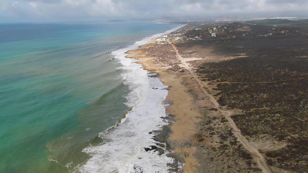 Oblique aerial view of Punta Gorda with pale surf curling onto sand and rocks, a long dirt road through brown-green vegetation, small distant buildings near the upper shoreline, and no visible people.