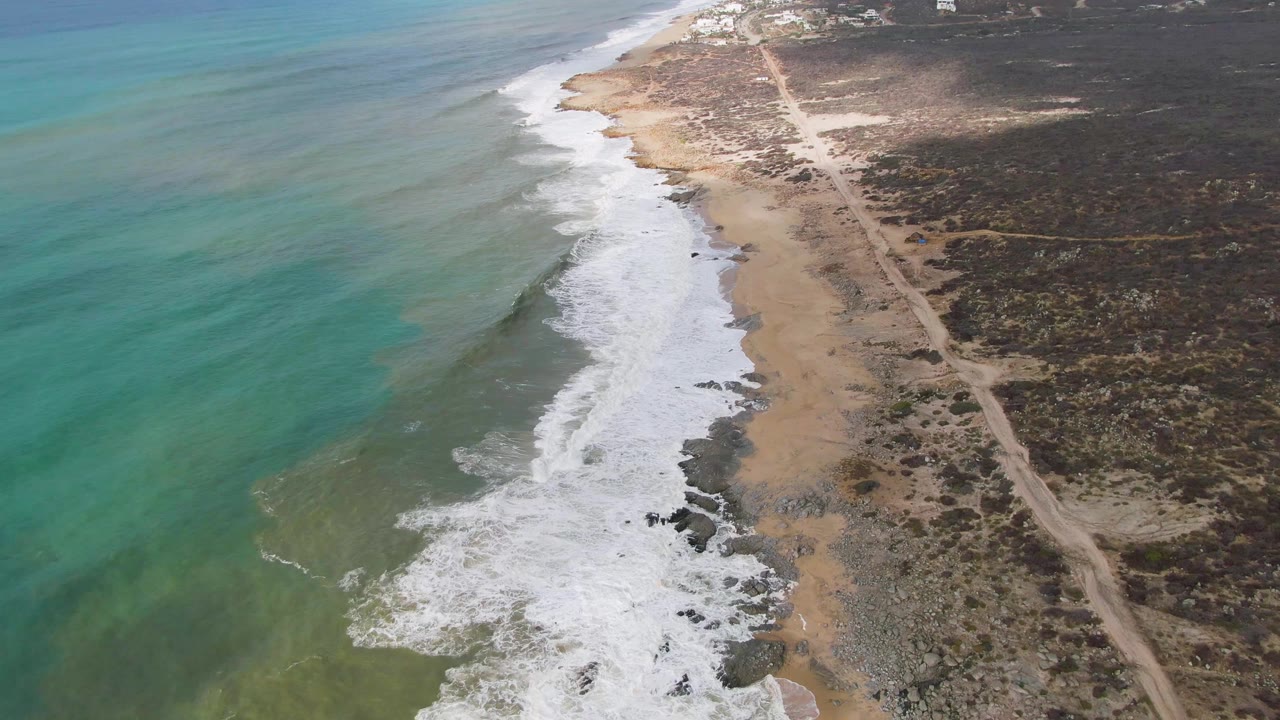 At 0:00, an aerial view over Punta Gorda shows turquoise water, white surf, a sandy shoreline, inland scrubland, and a dirt road running parallel to the beach with no visible people.