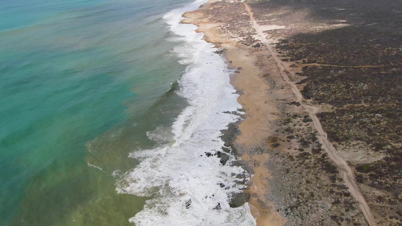 At 0:01, the aerial angle holds on Punta Gorda as white foam spreads farther along the shore, with green-blue water offshore, tan sand, dark rocks, dry vegetation, and no visible people.