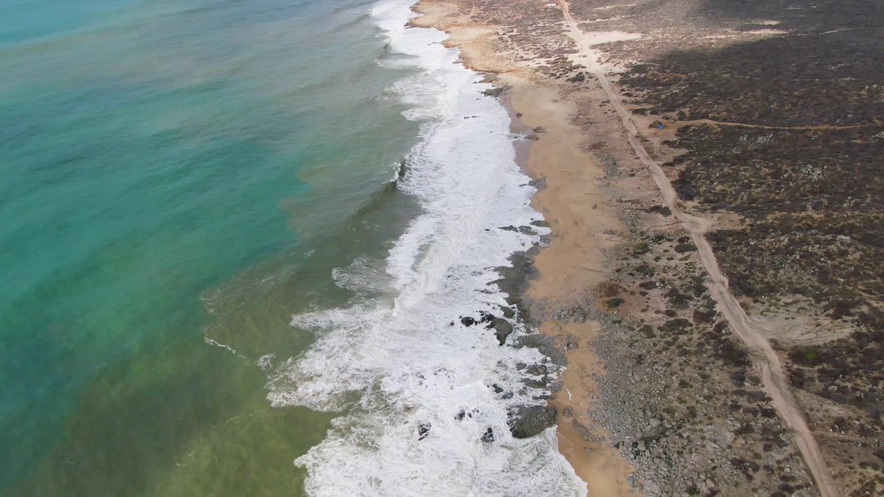 At 0:02, the Punta Gorda shoreline remains framed from above, with a broad band of white surf, visible dark rocks near the wash, dry brown scrubland, a straight dirt road, and no visible people.