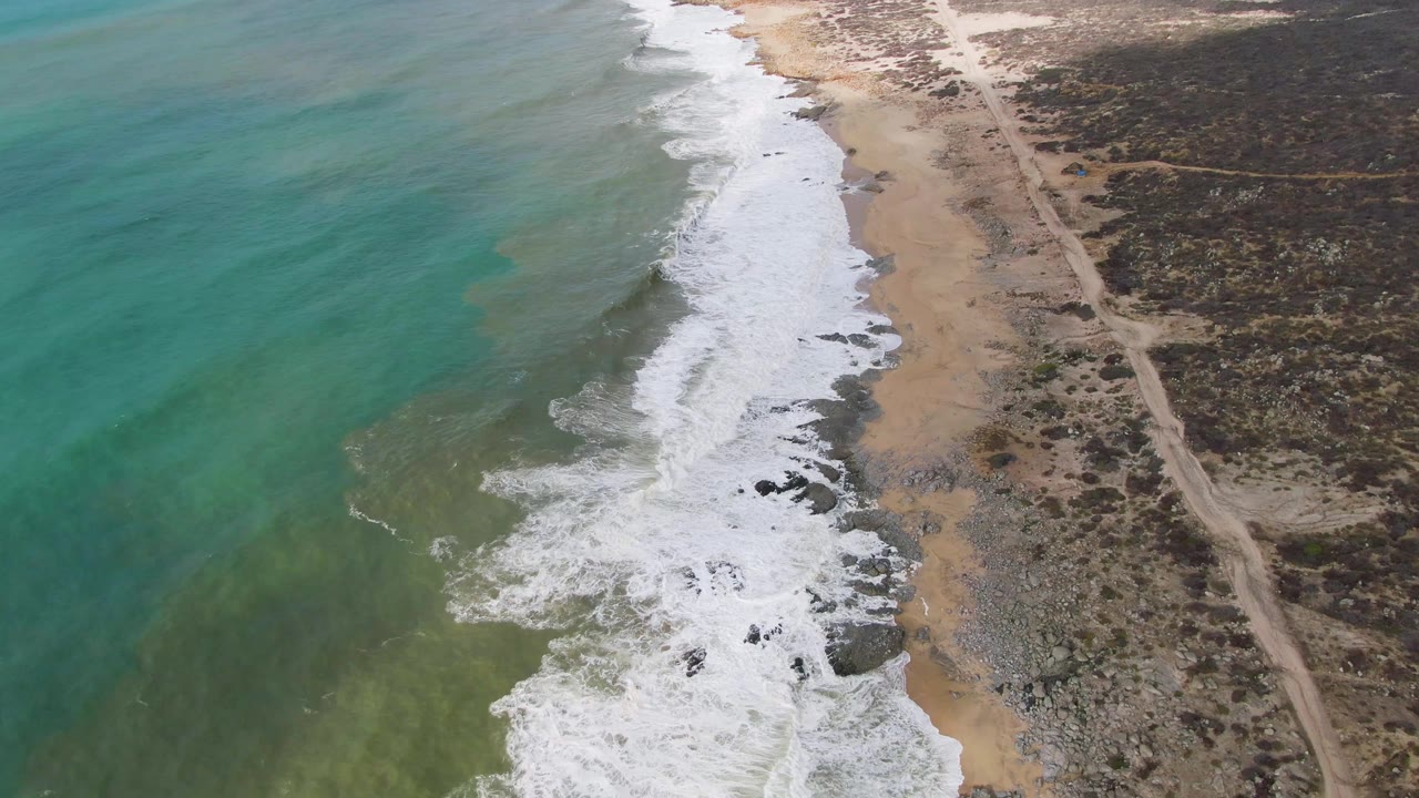 Aerial coastline with breaking waves, sandy beach, and scrubland road.