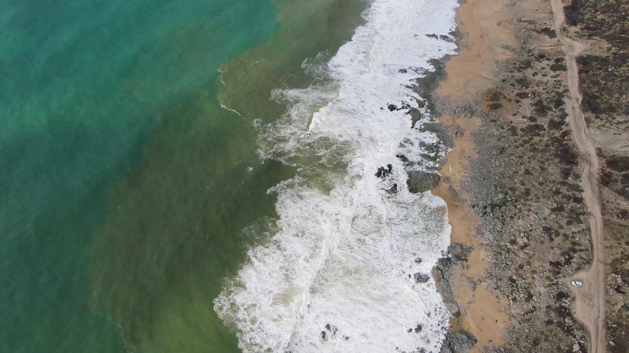 Overhead surf breaking along a rocky sand shoreline beside green water.