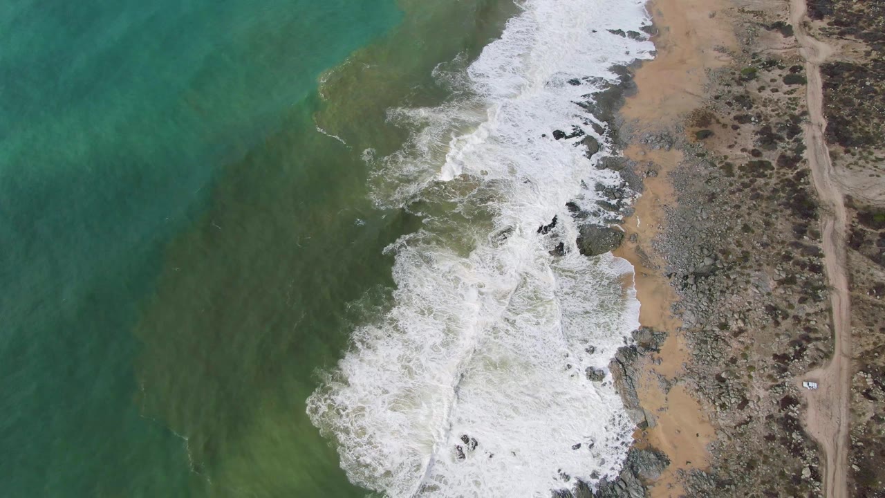 At 0:01, a broad patch of white foam rolls farther across the rocky sand beach at Punta Gorda East Cape, with green water offshore and dry scrubland beside an unpaved road, no visible people.