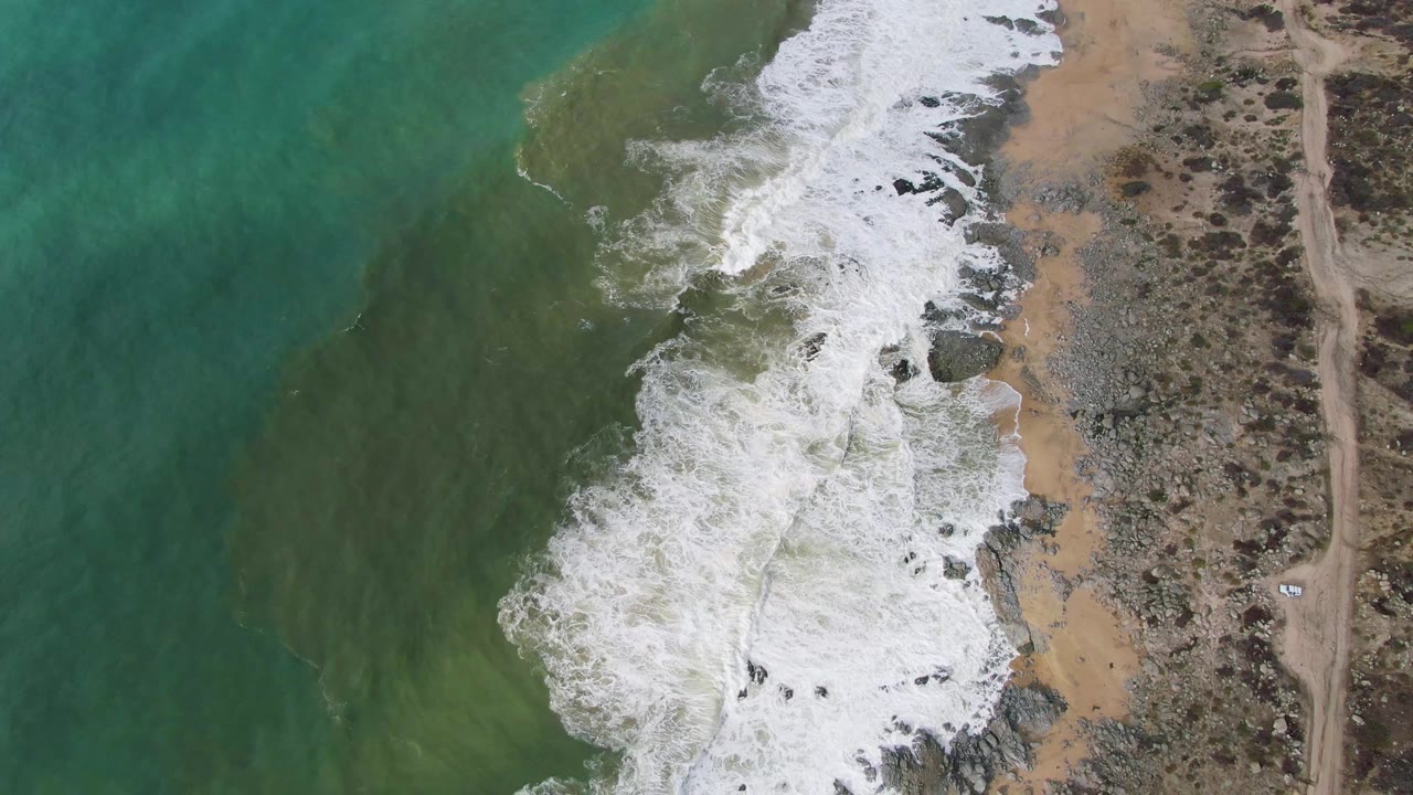 At 0:02, white surf curls diagonally into the Punta Gorda East Cape shoreline, with turquoise water to the left, tan sand and dark rocks at center, and scrubland with a dirt road to the right, no visible people.