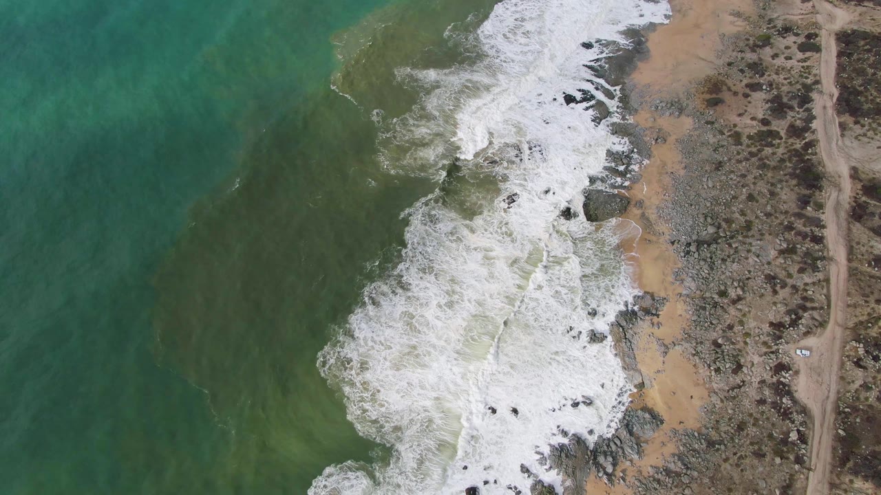 At 0:03, the overhead Punta Gorda East Cape view shows foamy wash covering much of the rocky shoreline while darker green water remains offshore and scrubland borders the beach, no visible people.