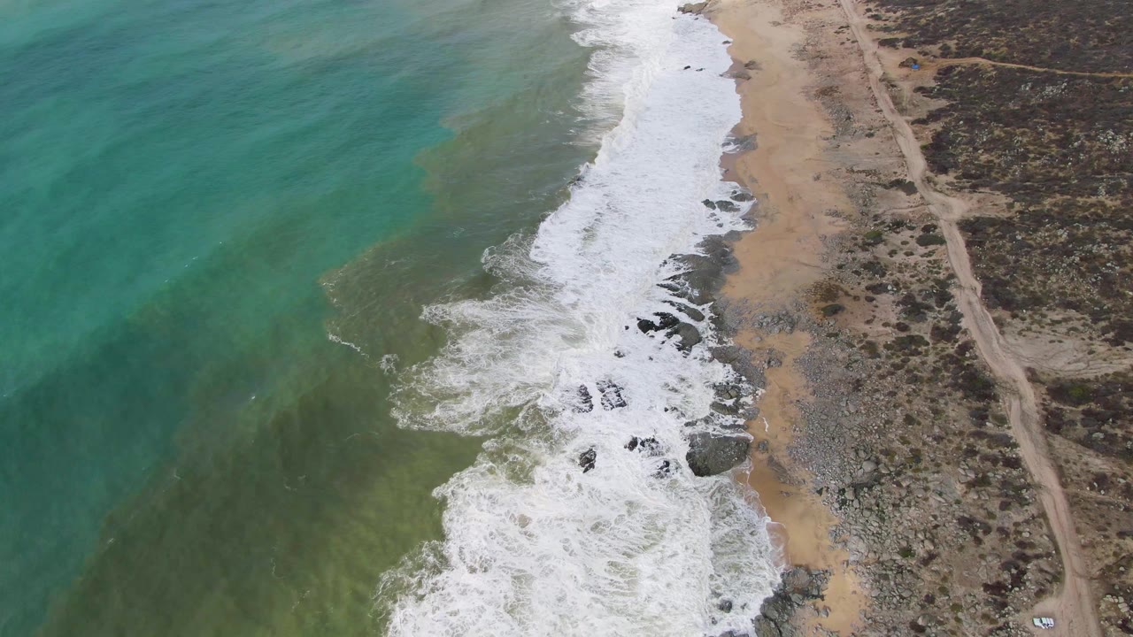 Aerial coastline with turquoise water, white surf, rocks, sand, and a dirt road.