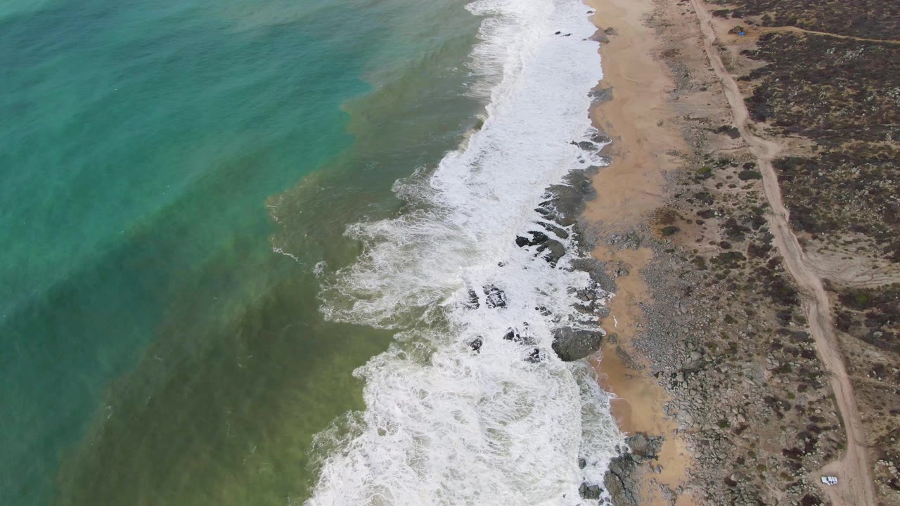 At 0:01, the Punta Gorda shoreline shows a larger band of white foam pushing across dark rocks and tan sand, with green-blue water offshore, scrubland to the right, and no visible people.