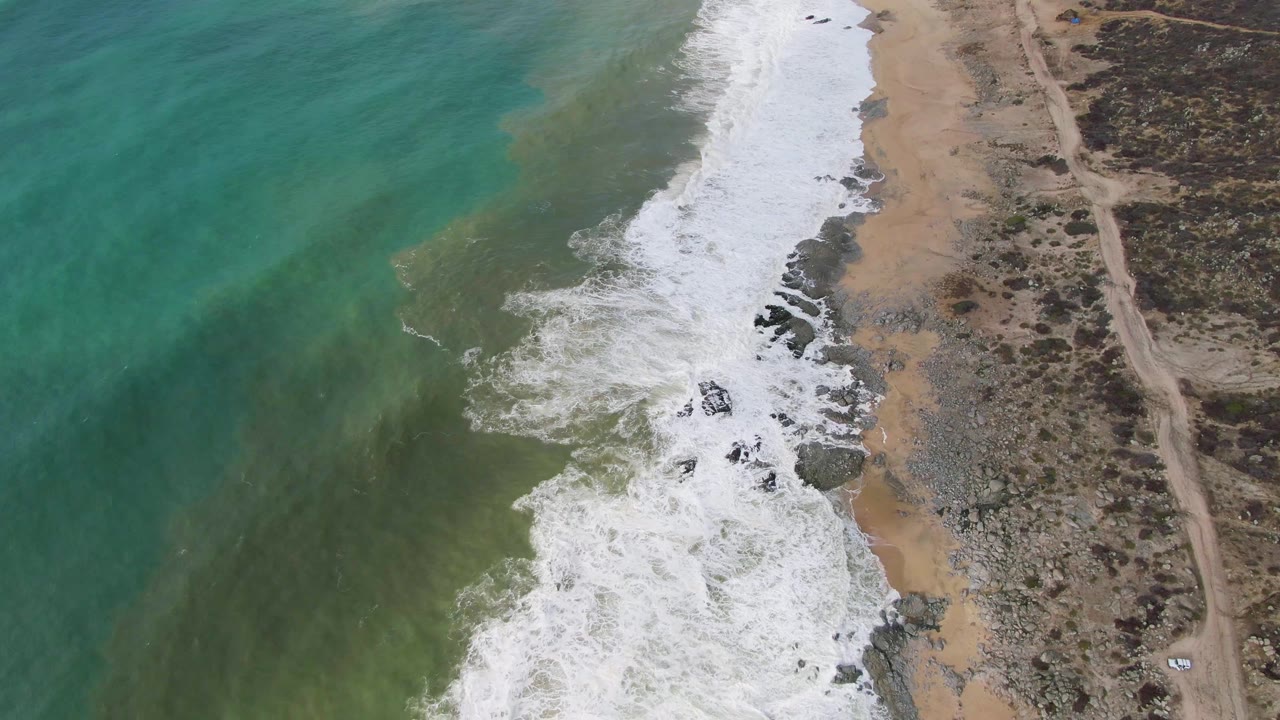 At 0:02, an aerial frame over Punta Gorda captures a fresh wave curling into the rocky beach, open turquoise water to the left, pale sand beside the surf, a dirt road inland, and no visible people.