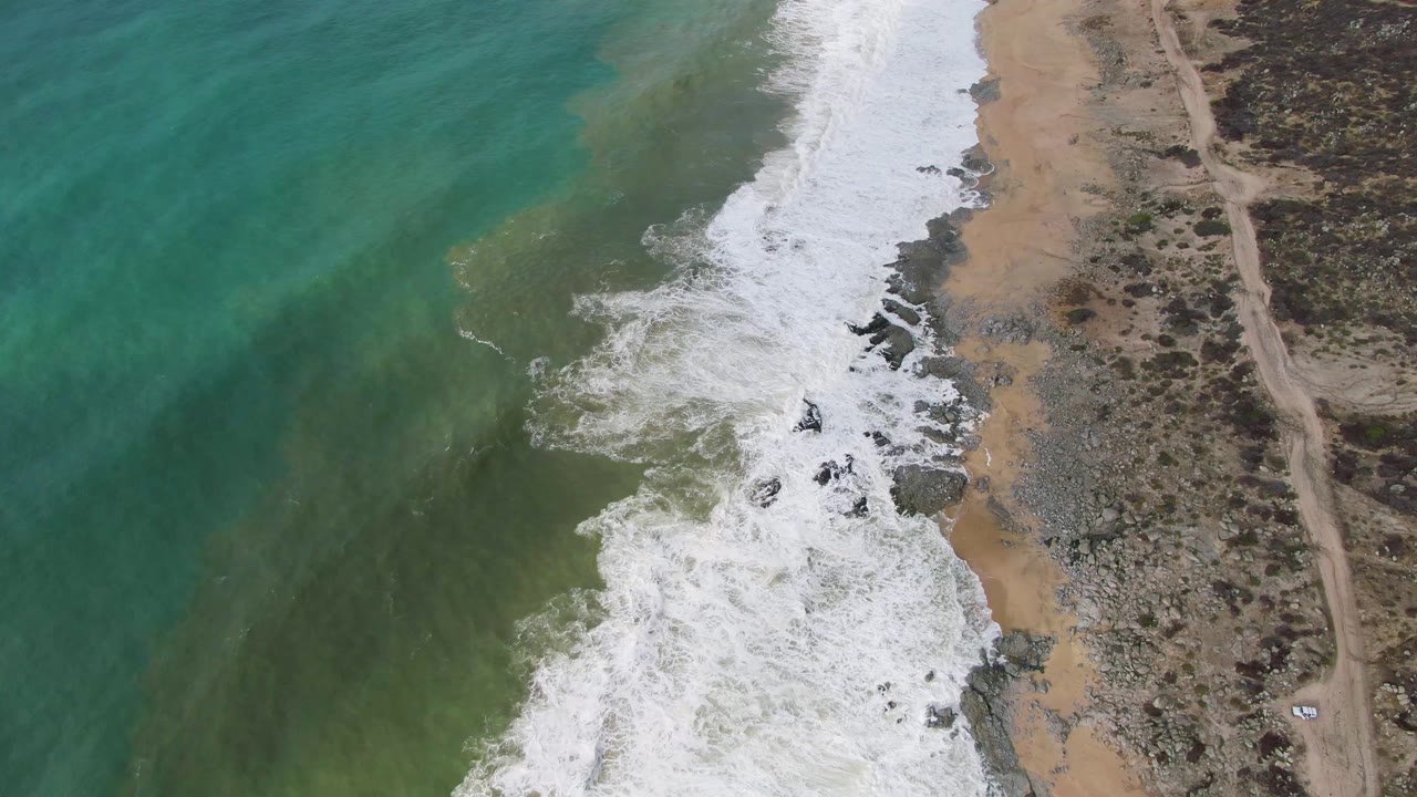 At 0:03, the Punta Gorda coast is seen from above as broad white surf fills the lower shoreline, rocks line the beach, turquoise water fades offshore, and the right side holds scrubland and a dirt road with no visible people.